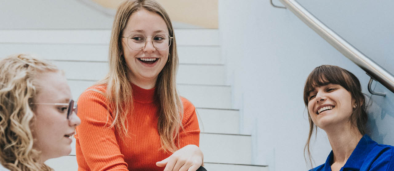 Een lachende studente uit de bachelor orthopedagogie zit samen met enkele medestudenten op de trap van gebouw T van HOGENT.