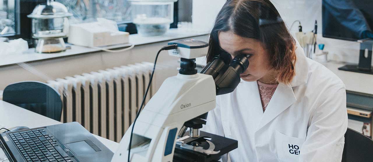 Een student uit de graduaatsopleiding laboratoriumassistentie aan HOGENT met een labojas kijkt door een microscoop.