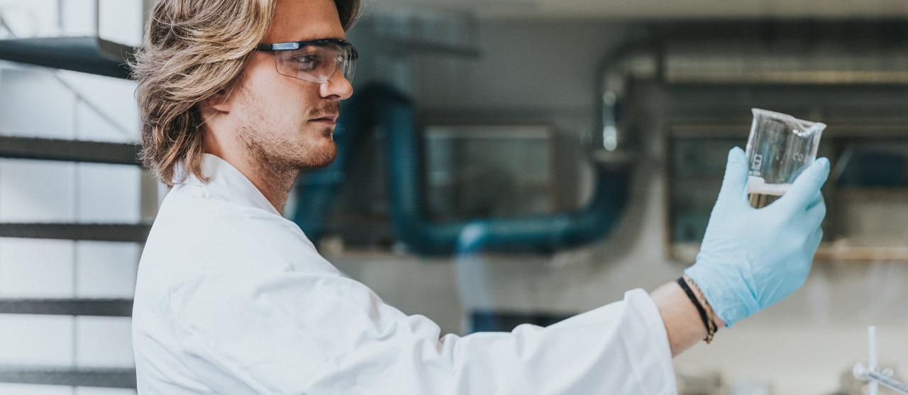Een student uit de graduaatsopleiding laboratoriumassistentie aan HOGENT met een labojas, handschoenen en een veiligheidsbril inspecteert een vloeistof in een glazen recipient.