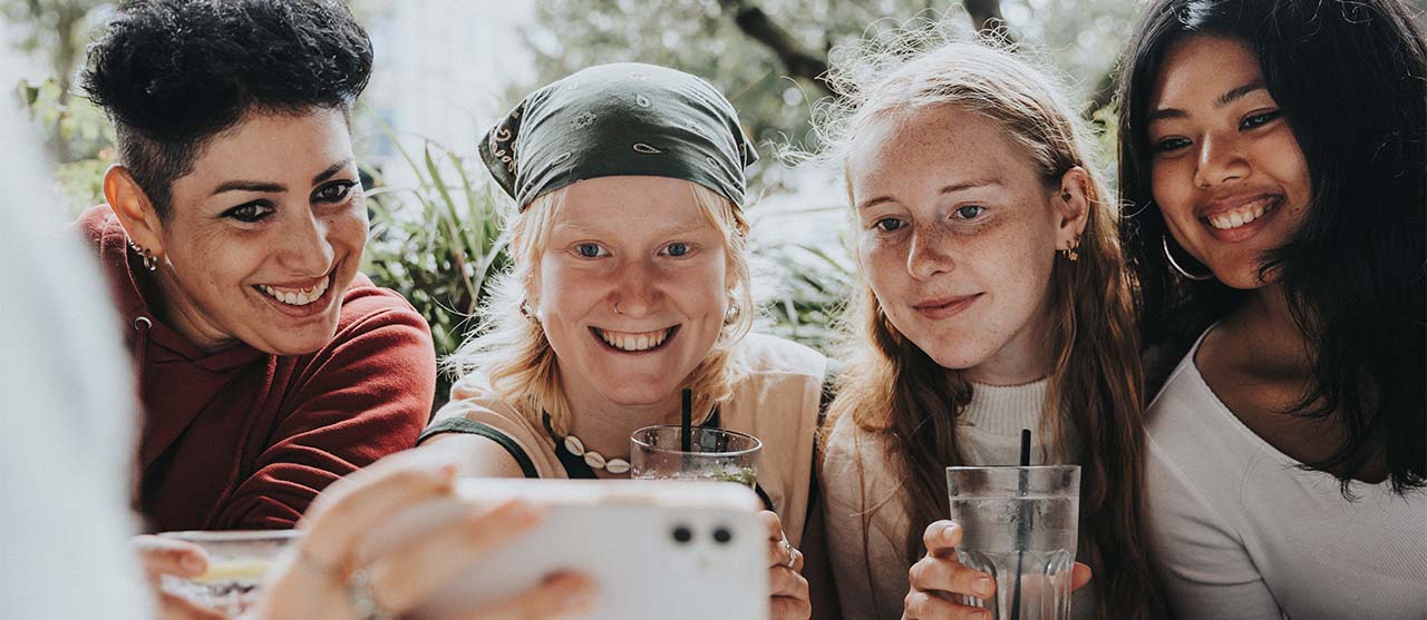 vier lachende studentes die kijken naar een gsm-scherm en een drankje vasthouden