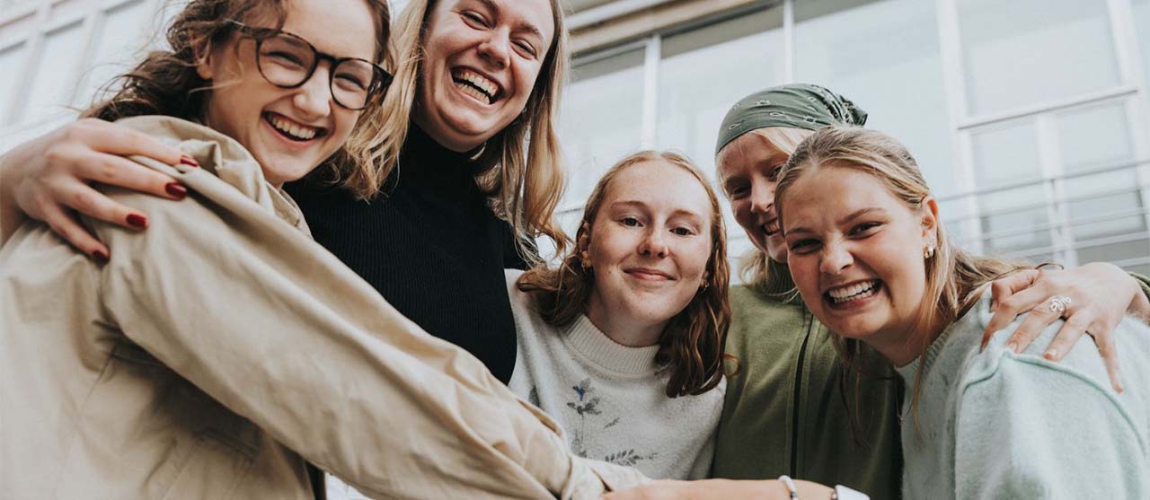 Vijf lachende studentes uit de bachelor sociaal werk van HOGENT omhelzen elkaar op de campus.