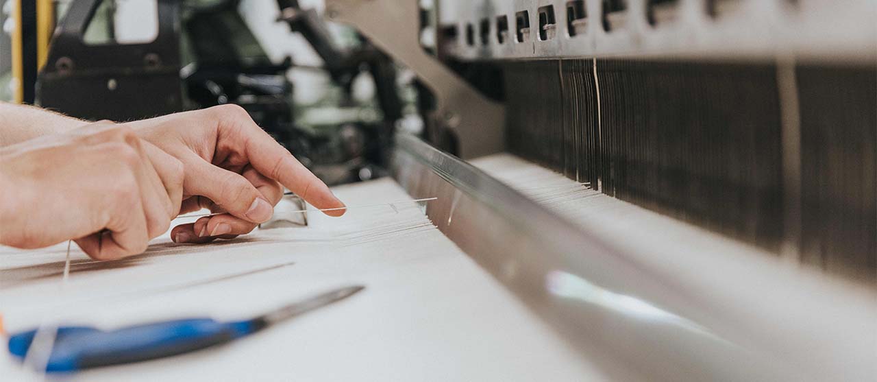Student textieltechnologie van HOGENT haalt kettingdraden door het riet van een industriële weefmachine.